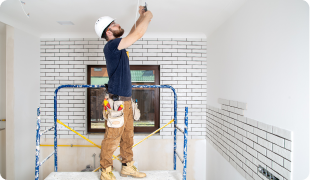 Construction worker renovating room, installing tiles on wall frame, ladder and tools nearby.